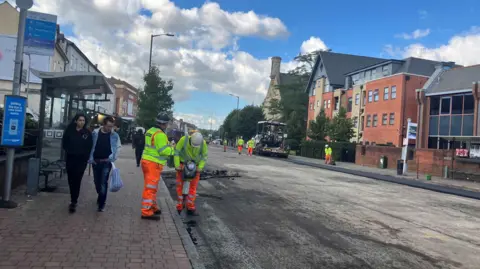 Highway workers can be seen in orange trousers and yellow jackets working on the resurfacing of a bus lane on the Oxford Road, in Reading.