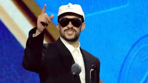Bad Bunny accepting a Grammy award. He is wearing a blacksuit with shite shirt, a apir of sunglasses and a white cap. 