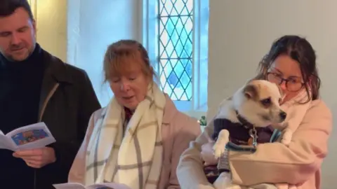 Luke Deal/BBC Two women and a man are stood taking part in a church service. One of them women is holding a white and brown dog in her arms. She is wearing a beige coat and glasses and is not looking at the camera. 
