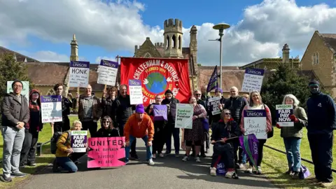 A group of around 20 people stand outside a historic building at a university campus on a partly cloudy day. They are on strike and are holding banners and placards calling for more pay. Most of them are wearing coats. They are standing on a path between two grass verges.