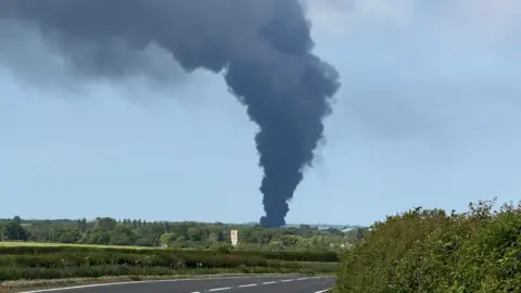 Steve Hubbard/BBC Dark grey smoke funneling out from base to top in the centre of the photo. There are fields with hedges around the smoke plume and a road in the foreground.