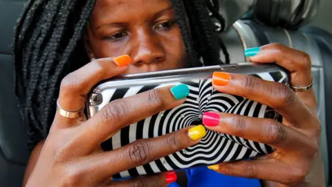 A woman with braids and multi-coloured nail varnish hold ups a mobile phone encased in a zebra design cover