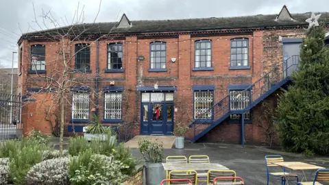 The exterior of the Emma Bridgwater gift shop which is made from red brick with windows and doors painted in blue trim. There is a Christmas wreath on the door and tables, chairs and plants can be seen in the foreground. 