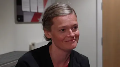 A woman sat down smiling wearing a black overshirt.