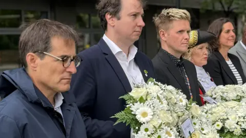 Living Bankside A group of people in formal attire stand outdoors holding wreaths and bouquets of flowers, including white and red arrangements with ribbon, during a ceremonial gathering.