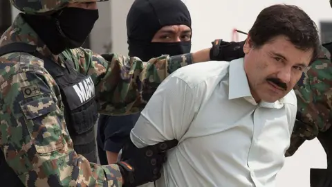 Getty Images Membros da Marinha mexicana, usando balaclavas, escoltar El Chapo após sua prisão. El Chapo tem um bigode e está olhando para a câmera enquanto um dos fuzileiros navais o segura pelo pescoço e pelo braço.
