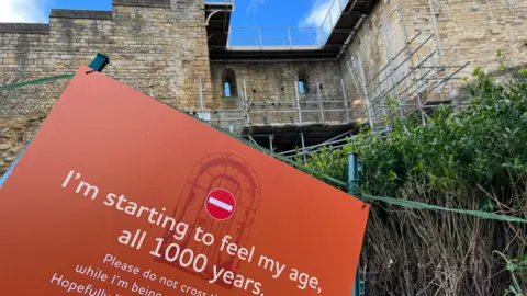 Scaffolding covers the west gate of the castle. In the foreground is a sign reading "I'm starting to feel my age, all 1000 years"