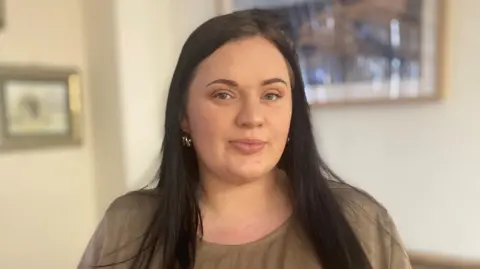 Fiona Goddard, who has long dark hair and wears a brown top, looks straight at the camera.