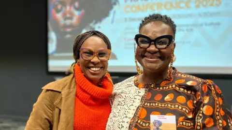 Jon Wright/BBC Chez Eradiri (left) and Revd Valerie Lolomari stand smiling at the camera. There is a big projector screen behind.