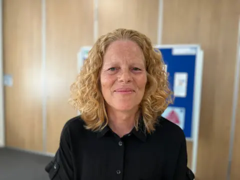 A woman with shoulder-length curly hair is sat in a boardroom with a noticeboard and light-coloured walls softly out of focus in the background.
