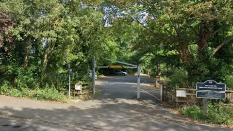Google The entrance to a car park, with a height barrier, trees on both sides and a sign saying welcome to Radford Road car park