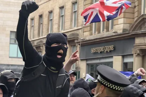 BBC A masked individual in dark clothing raises a clenched fist while standing close to a police officer in a high-visibility jacket and checkered hat, with a crowd gathered behind and a Union Jack flag visible above on a city street lined with stone buildings and shopfront signage reading “Sostrene Grene.”