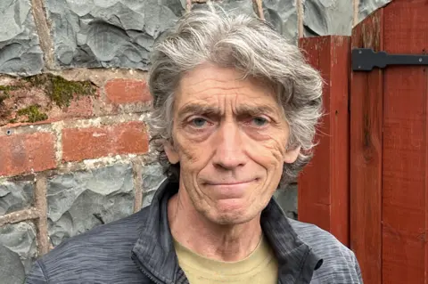 Ian Young is standing in front of a wall made of bricks and stone, as well as a large red wooden gate. He has grey hair and is smiling at the camera.