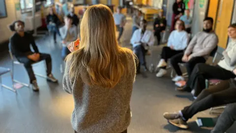 BBC A group of young people who are not in education, employment or training sit in a semicircle during a group coaching session on employability skills. 