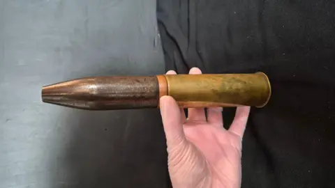 Kirkgate Arts and Heritage A brass WW2 artillery shell. The shell is being held by a hand which is in shot. The shell is about 15-inches in size. 