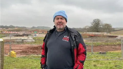 A man is standing in front of a fenced-off field with HS2 construction in the background. He is wearing a navy T-shirt that says "Stop HS2". He is wearing a light blue beanie and has his hands in his pockets.