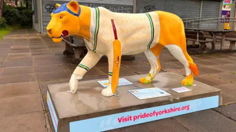 A decorative lion sculpture displayed outdoors on a paved area. The lion is painted in bright shades of yellow and cream and is designed to look as if it is wearing a knitted jumper, with detailed, repeating stitch-like patterns covering its body in white and pale yellow tones. A cricket bat is painted along the side of the lion, adding a sports-related theme to the design. The lion is positioned mid-step, with one front leg lifted, giving the impression of movement.