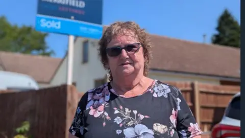 Viv Matthews poses in front of her home. She is wearing a floral dress and has short, curly hair and dark sunglasses. A 'sold' house sign can be seen behind her