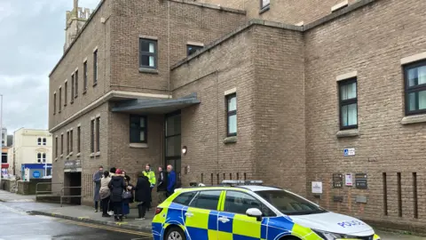 A group of people stood outside of the building, with police car nearby. The building has brown bricks and is near other shops.