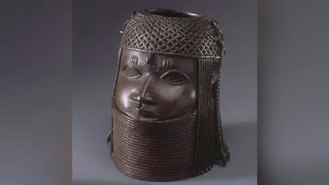 Commemorative head of the Oba, or king. It is a dark brown metal sculpture showing a face with a chain-mail helmet on its head and a metal collar up to its chin.