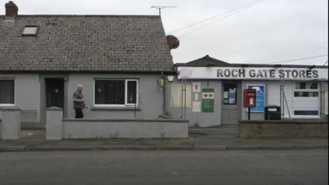 Mr Brentnall walking towards the grey concrete building which has a faded Post Office sign next to text saying Roch Gate Stores.