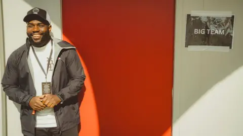 Big Team CIC Javeon McCarthy is smiling next to a festival cabin, which has red door with a sign that reads Big Team. Javeon is wearing a black jacket and a black cap with a white t-shirt 
