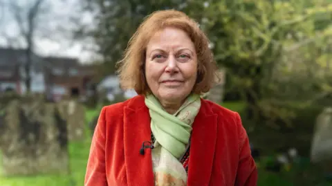 Ian Palmer/ BBC A woman with short red hair looks directly into the camera. She is wearing an orange velvet jacket and green patterned scarf. She is standing in a graveyard and graves can be seen in the background. 