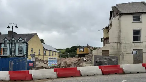 A large digger is pictured over the rubble of a building. A large gap is visible between two exterior buildings and a number of cordons are in place