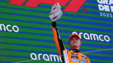 PA Media Lando Norris is holding up his trophy in front of a green and blue screen. He is wearing an orange kit and cap.