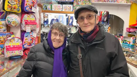 Patricia Ingle - a woman with a long purple scarf around her head, glasses and short grey hair - is smiling into the camera in a black coat. Next to her is Alan Ingle - a man with a black cap on, a Sprayway black coat and a colourful scarf. Behind them are children's colourful backpacks and toys in a shop.