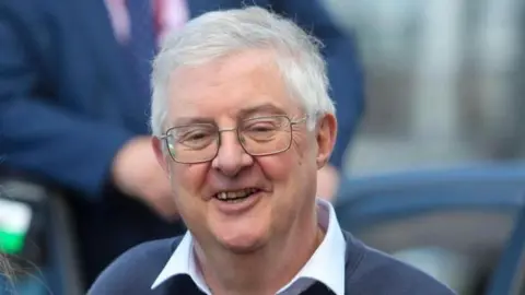 A head and shoulders picture of Mark Drakeford smiling. He is wearing spectacles, a white shirt and blue jumper