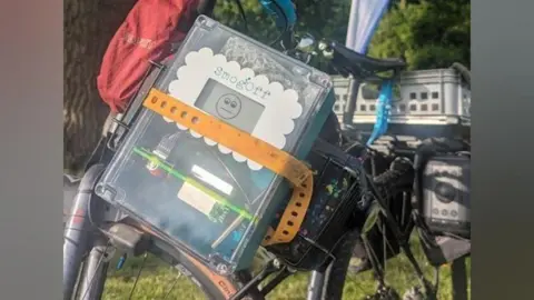 Al Pagan A clear plastic device with a screen and electric circuitry, with the word SmogOff and a smiley face on the screen. The device is attached to a bicycle.