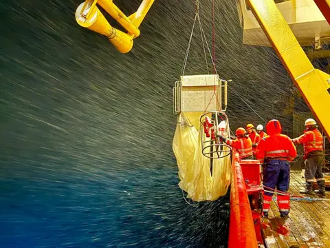 Prof Daniel J Mayor @oceanplankton Five scientists wearing high-vis orange jackets and dark trousers on board the Sir David Attenborough polar ship. They are working with a fishing net equipped with 9 closing nets and has a 1 × 1 m mouth. There is a large yellow crane above them.