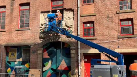 BBC/Steve Jones A workman is in a cherry picker in front of the frieze which has been partially removed