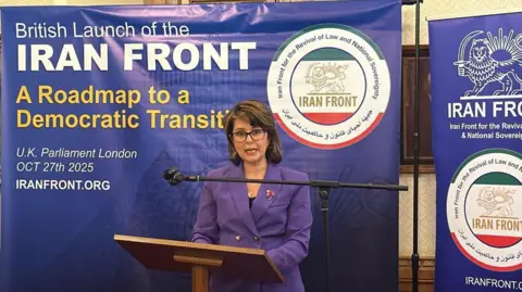 Mattie Heaven is wearing a purple suit and addressing a news conference from a lectern. She is standing in front of a banner reading 'British Launch of the Iran Front; A Roadmap to a Democratic Transition', held at the Palace of Westminster in October 2025.