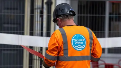 A Thames Water employee wearing a hi-vis uniform reads a document inside a red folder while on a site