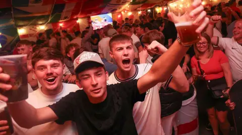 England supporters cheer after their team's first goal as they watch the television broadcast of the 2026 World Cup Group L football match between England and Panama, at The Clock pub in Hebburn, north-east England. The scene has a large crowd with three men in the front celebrating with their arms up. 