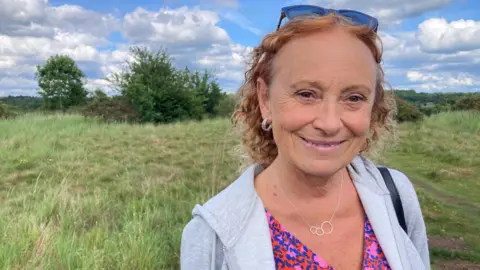 A woman wearing a grey jumper, smiling, stood in front of a large grassy field.