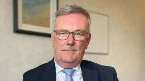 PA A white haired man with grey glasses wearing a dark blue suit with a white shirt and light blue tie with pink polka dots sitting in front of wall with cream wallpaper and paintings. 