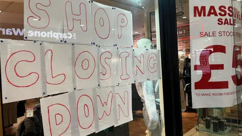 Sign in shop window which says 'shop closing down'. Clothes are visible through the window. 