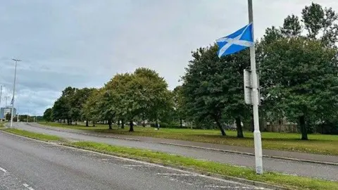 A blue and white saltire flag is fluttering from a lamppost on an Aberdeen street, lined with trees.