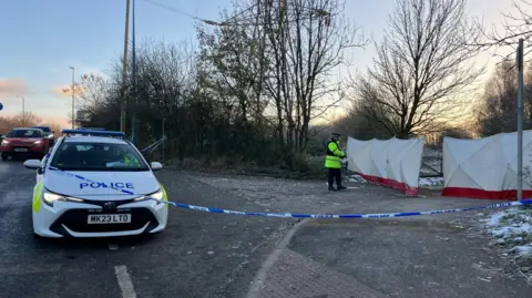 BBC/ Matt Trewern A police car at the scene in Salford, with police tape and a lone police officer standing in front of white screens which appear to be about 7ft (2m) tall.