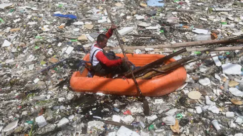 A worker conducts clearing operations to collect floating trash and debris that have accumulated along a water channel following successive days of monsoon rains