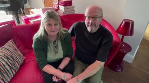 Jeanette and Craig are sat on a red sofa. Jeanette is wearing a green top and cardigan and dark trousers. Crag is wearing dark rimmed glasses, a black t-shirt and green trousers.