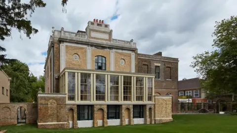 Getty Images A restored early 19th century English country house on three floors stands in front of a lawn. Newer residential buildings are visible in the background. 