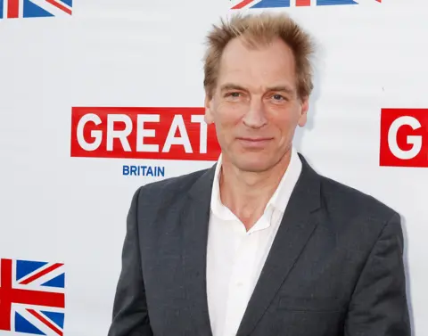 Reuters Actor Julian Sands - a blond man in a grey suit and white shirt - stands in front of a sign which reads Great Britain. 