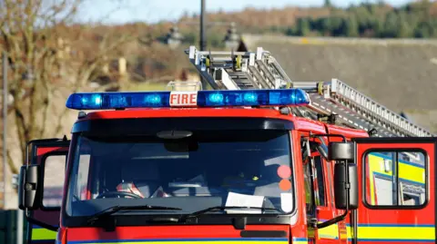 The top half of a red fire engine. The vehicle is red with high visibility stripes. There are blue lights on the top along with ladders. Doors on both sides are open. The front of the vehicle is empty. 