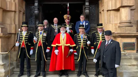 The town crier in a long red cloak is standing on steps next to four men in long black tail coats with gold detailing across their chest and top hat. They are wearing white gloves, holding batons and some have medals pinned on them. There is a man in a black three-piece suit and top hat stood in front of them, and four people in various suits and uniforms stood behind.