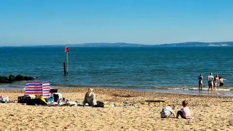 Penny Wade A beach scene with some people sat on the sand and a few others venturing out into the water. The blue sky is clear with hills in the background beyond the water.