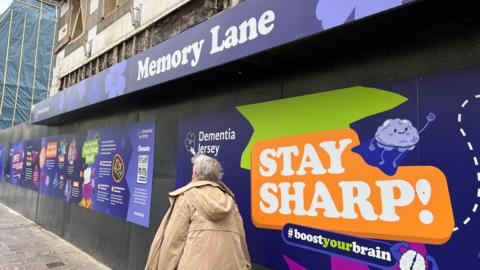 A woman walks past the artwork hoardings with the sign Memory Lane above her
